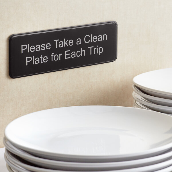 A stack of Tablecraft plastic plates with a sign that says "Please Take a Clean Plate for Each Trip" on a table.