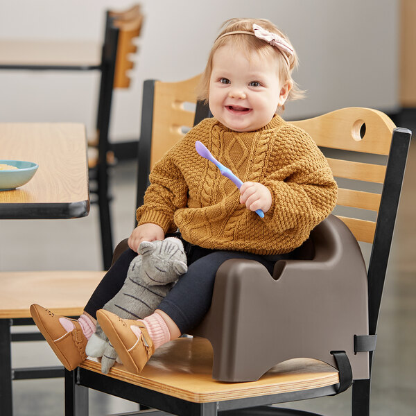 A baby sitting in a Carlisle brown booster seat holding a spoon.