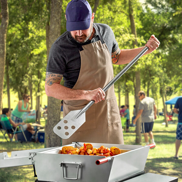 A man in an apron using a Fourt&#233; perforated stainless steel paddle to cook food on a grill.
