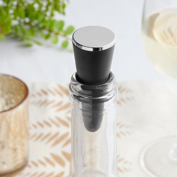 A close-up of a bottle with a Franmara silver-plated plastic stopper with a black and silver top next to a glass on a table.