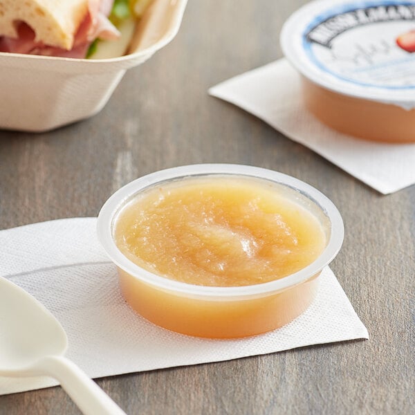 A plastic container of Musselman's unsweetened applesauce on a napkin next to a sandwich.