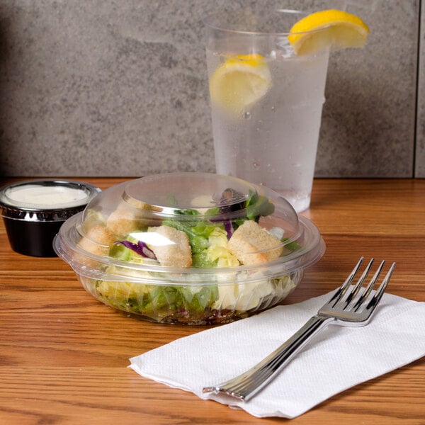 A Dart PresentaBowls clear plastic container filled with salad on a table next to a glass of water.