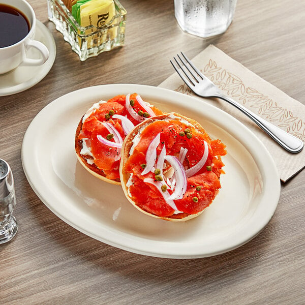 An Acopa narrow rim oval stoneware platter with a bagel and two sandwiches on it on a table with a cup of coffee.