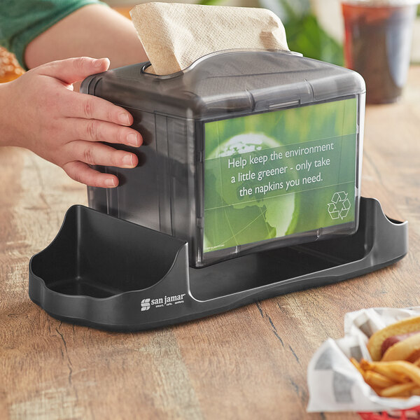 A hand holding a San Jamar napkin dispenser filled with interfold napkins on a counter.