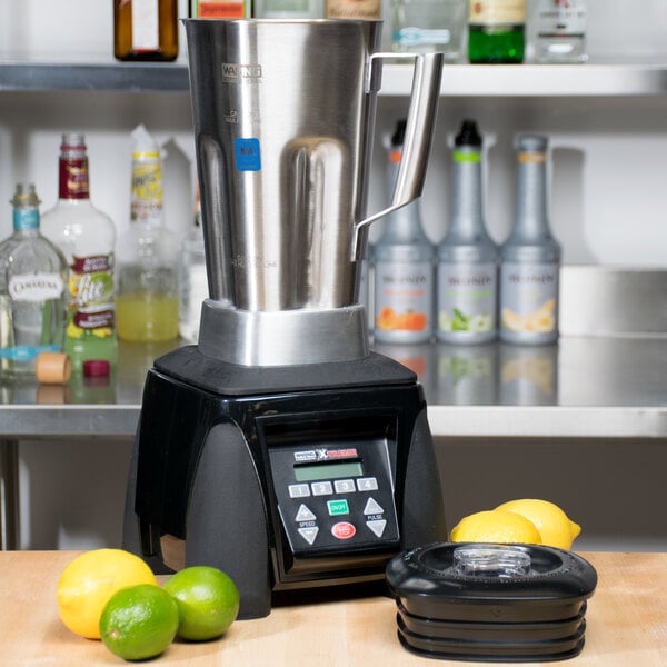 A Waring commercial blender with a stainless steel container on a counter with lemons and limes.