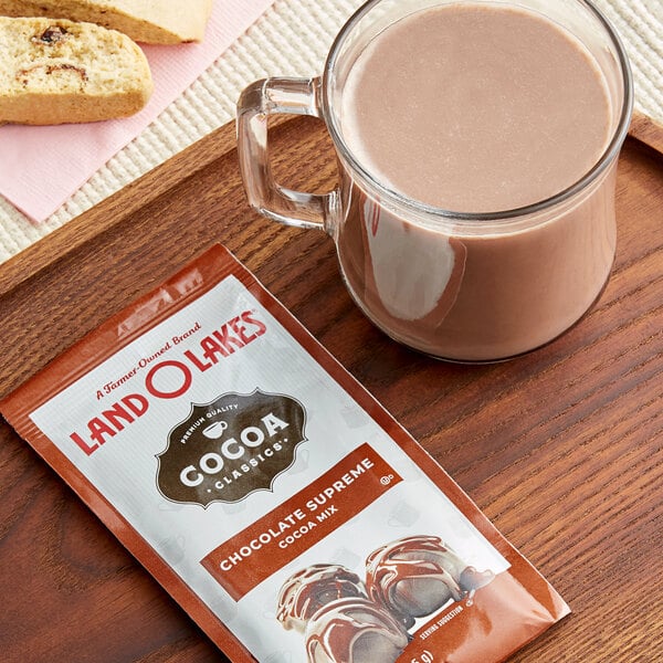 A glass mug of hot chocolate next to a Land O Lakes Cocoa Classics Chocolate Supreme cocoa mix packet.