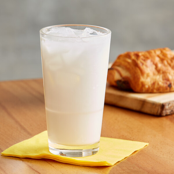 A glass of white Land O Lakes Arctic White Chocolate cocoa with ice and a croissant on a wooden surface.