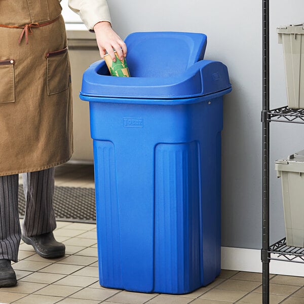 A man wearing a brown coat and striped pants standing next to a blue Toter Slimline commercial trash can.