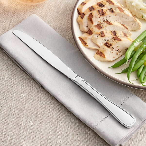 An Acopa stainless steel dinner knife on a napkin next to a plate of chicken and vegetables.