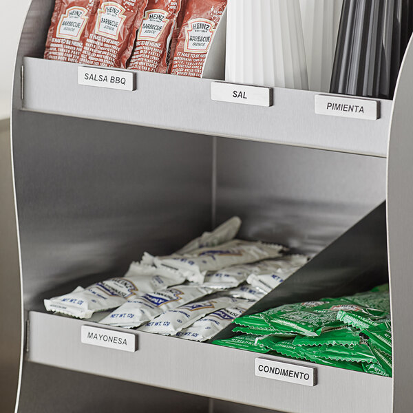 A silver metal shelf with magnetic stainless steel condiment labels in Spanish on a counter with condiments.