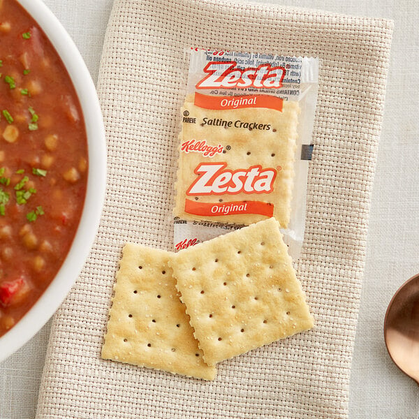 A bowl of Zesta saltine crackers next to a bowl of soup on a napkin with a spoon.
