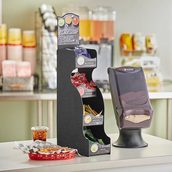 A ServSense black vertical countertop condiment organizer with 6 sections and header decals holding a variety of snacks.