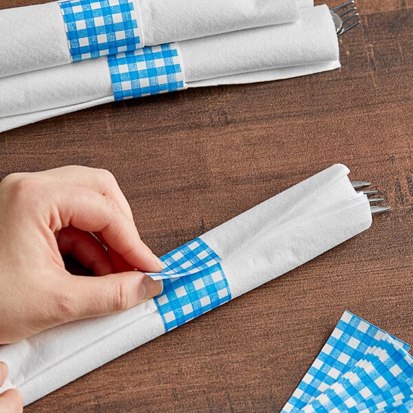 A hand using blue gingham checkered self-adhering paper napkin band to put a napkin on a table.