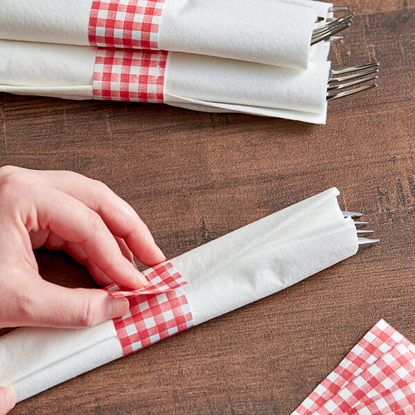 A person's hand using a red and white checkered paper napkin band to put a napkin on a table.