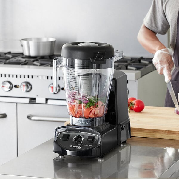 A man using a Vitamix XL blender to blend vegetables in a white bowl on a kitchen counter.