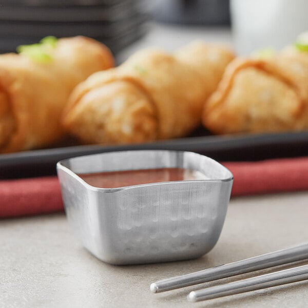 A close up of American Metalcraft stainless steel sauce cups on a tray with brown sauce.