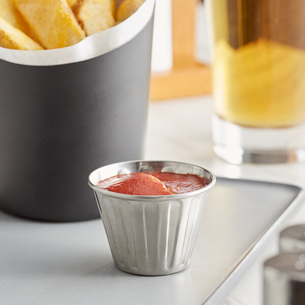 An American Metalcraft fluted stainless steel sauce cup filled with red sauce on a table with a pile of french fries.