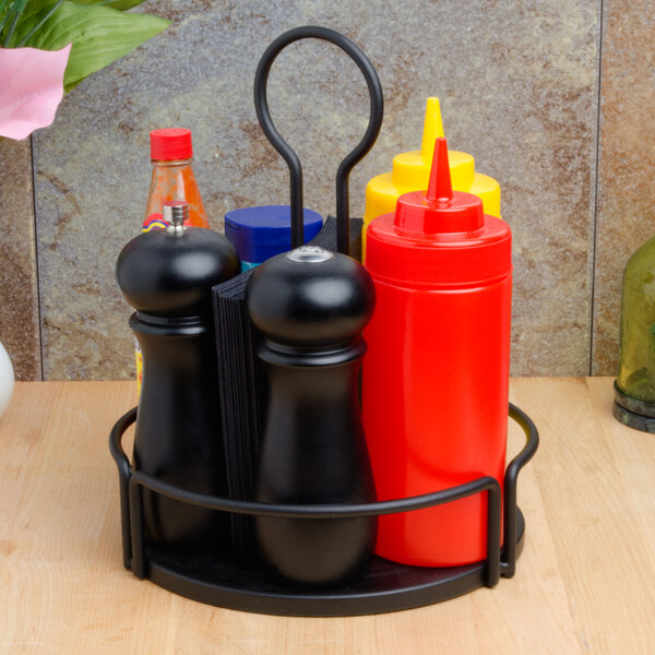 A Tablecraft black metal condiment caddy on a table with three condiments and a ketchup bottle.