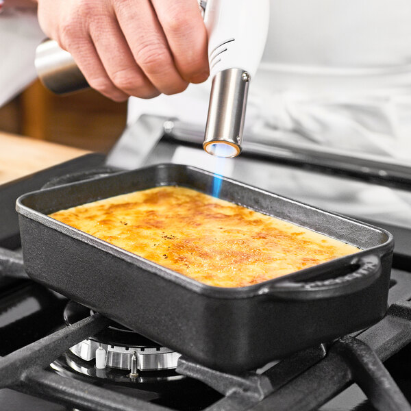 A person using a gas burner to cook a dish in a Chasseur black enameled cast iron rectangular casserole dish.