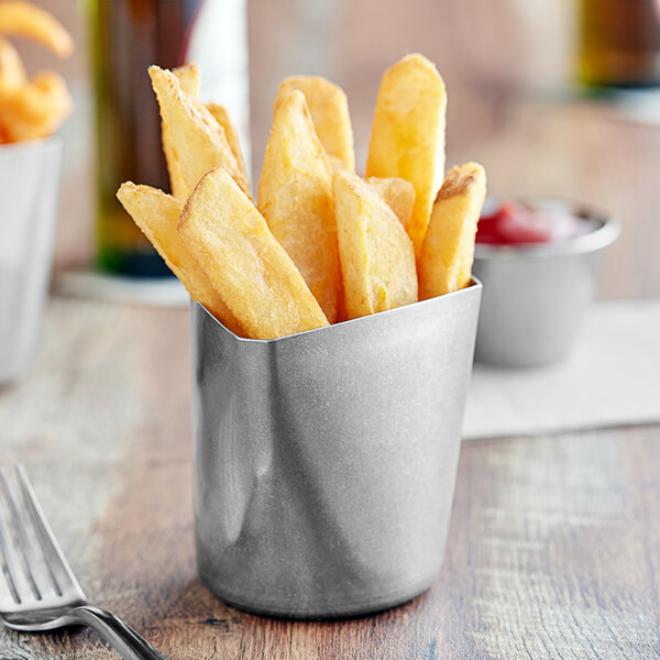 A Tablecraft stainless steel triangular fry cup filled with French fries on a table.
