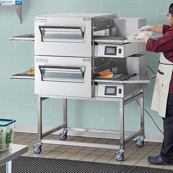 A man in a white apron and hat using a Lincoln Impinger II double conveyor oven.