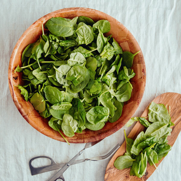 A Thunder Group woven wood salad bowl filled with spinach leaves on a wooden board.