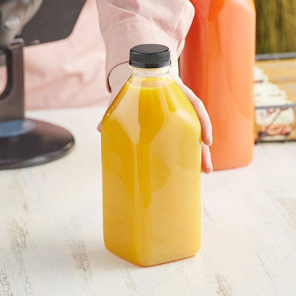 A hand holding a 32 oz. Milkman Square PET clear bottle with black lid full of orange juice.