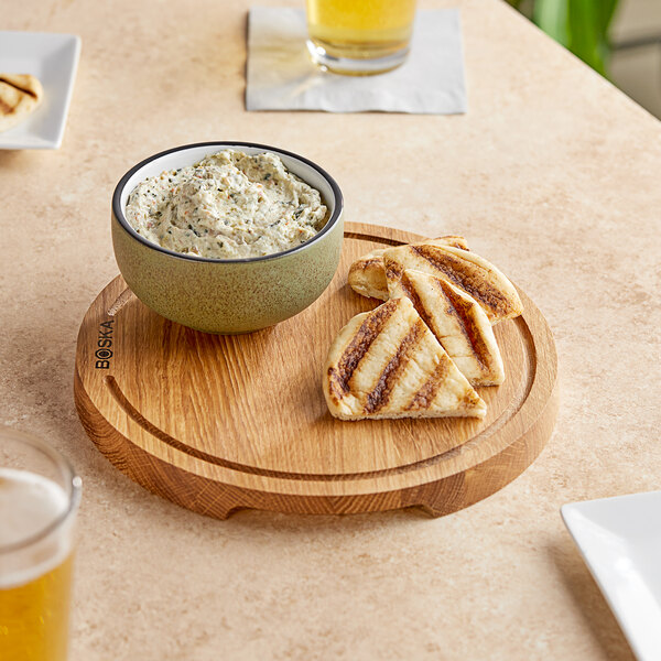A bowl of dip and grilled pita bread on a Boska small round European oak serving board.