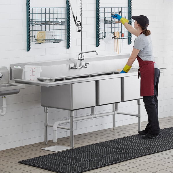 A woman in a black apron washing dishes in a Regency 3 compartment sink in a professional kitchen.