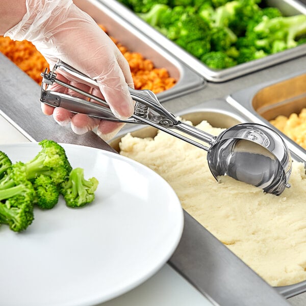 A person using a Choice stainless steel scoop to serve mashed potatoes over broccoli.