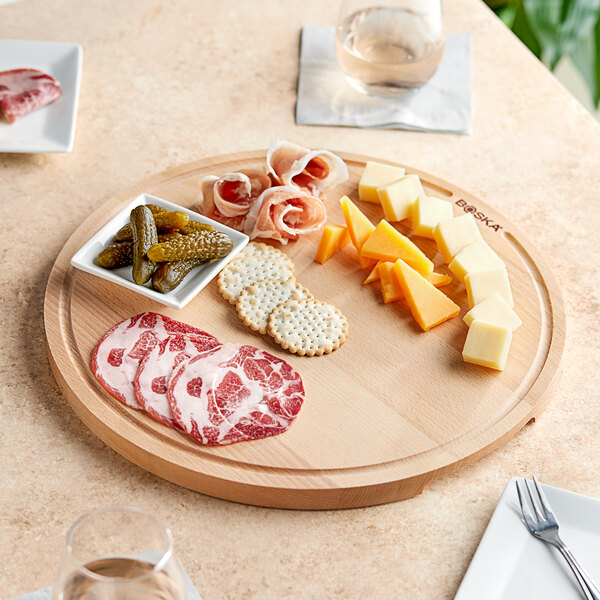 A Boska large round beech wood serving board with different types of meat and cheese on a table.