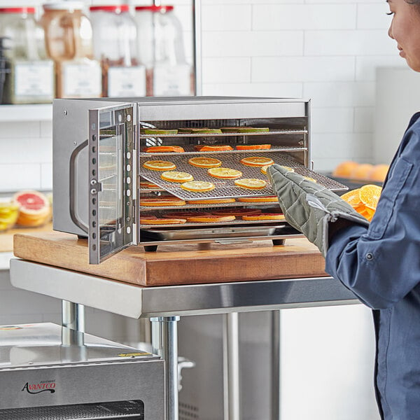 A woman wearing gloves places oranges in an Avantco stainless steel food dehydrator.