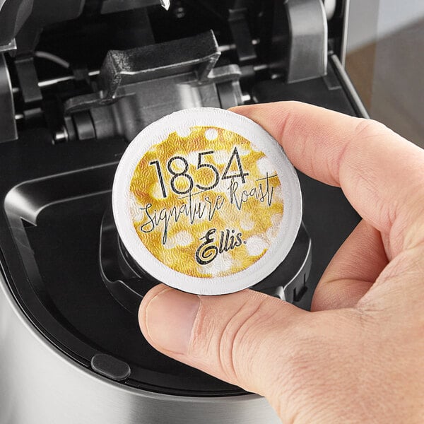 A hand holding a yellow and white box of Ellis 1854 Roast Coffee Single Serve Cups with black text.