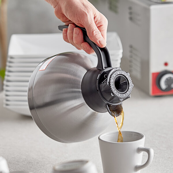 A person using a Thermos stainless steel coffee carafe to pour brown coffee into a white mug.