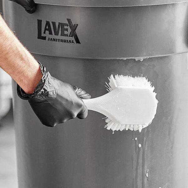 A hand in a black glove using a Lavex white floating utility brush to clean a trash can.