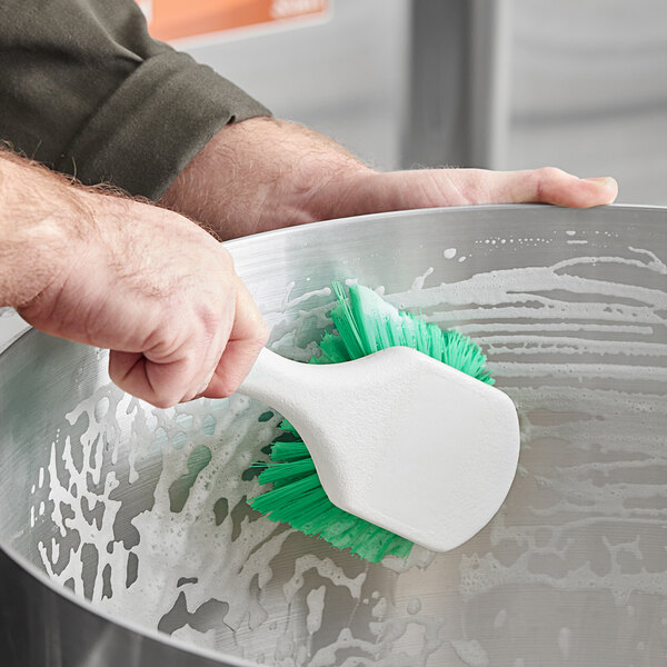 A hand using a Lavex green utility brush to clean a metal bowl on a counter.