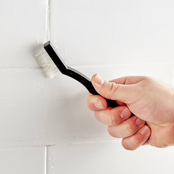 A person using a Lavex toothbrush style grout brush to clean a white tile wall.