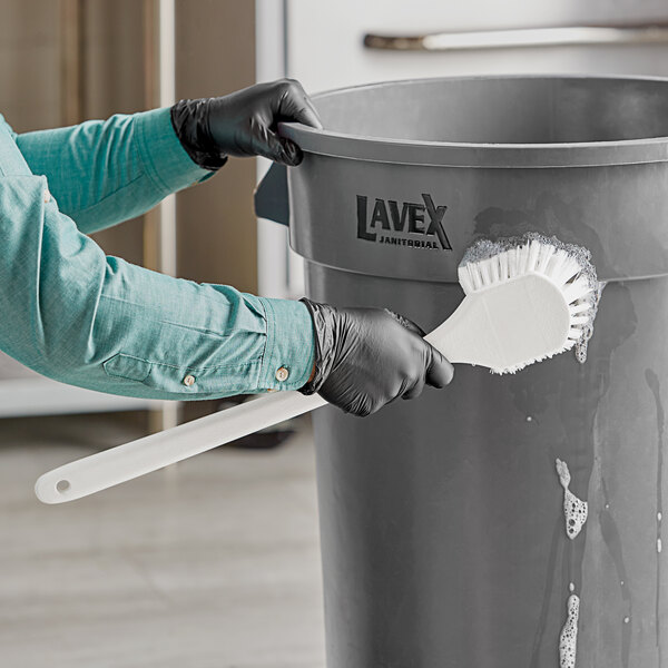 A person wearing black gloves using a white Lavex pot scrub brush to clean a black bucket.