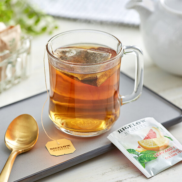 A glass mug of Bigelow tea with a tea bag and a white teapot on a tray.