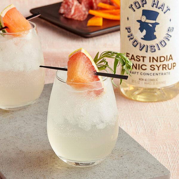 A glass of Top Hat Provisions East India Tonic with a slice of orange and a garnish on the rim.