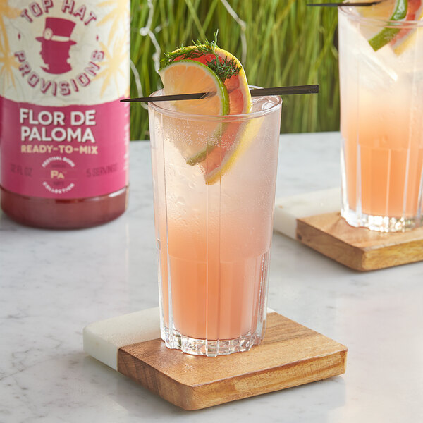 A glass of Top Hat Provisions Flor De Paloma Mix with orange slices and a straw on a wooden coaster.