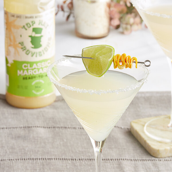 A bottle of Top Hat Provisions Classic Margarita Mix on a table with a glass of white liquid and a lime on top.
