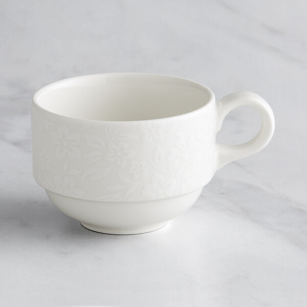 A close up of a white RAK Porcelain teacup with an ivory floral design and a handle.