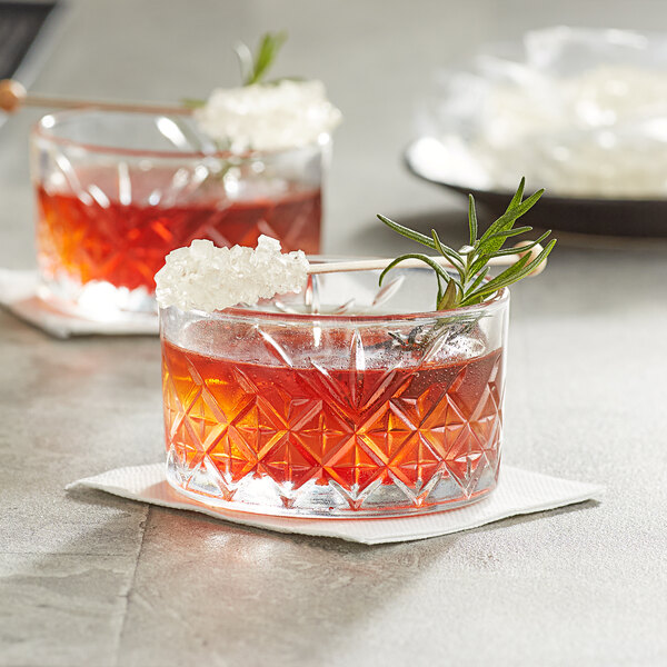 Two glasses of red liquid with Roses Dryden and Palmer White Wrapped Rock Candy Swizzle Sticks and rosemary sprigs.