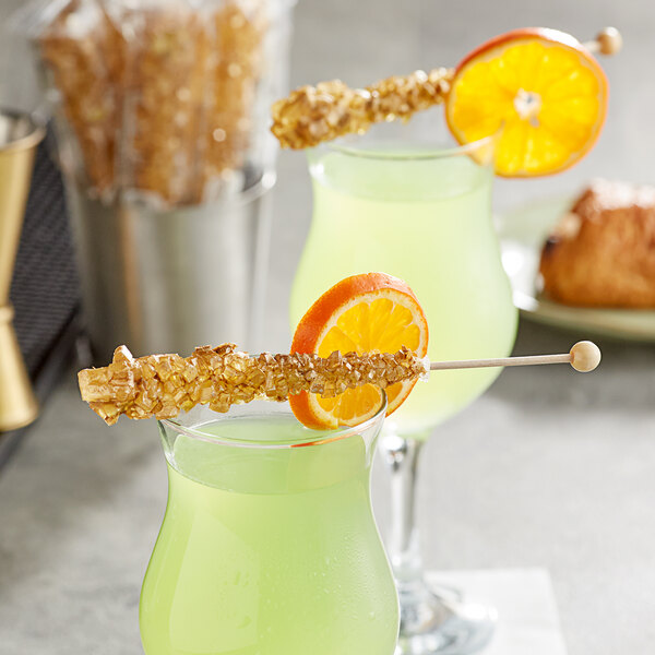 Two glasses of green liquid with Roses Dryden and Palmer gold wrapped rock candy swizzle sticks and orange slices on top.