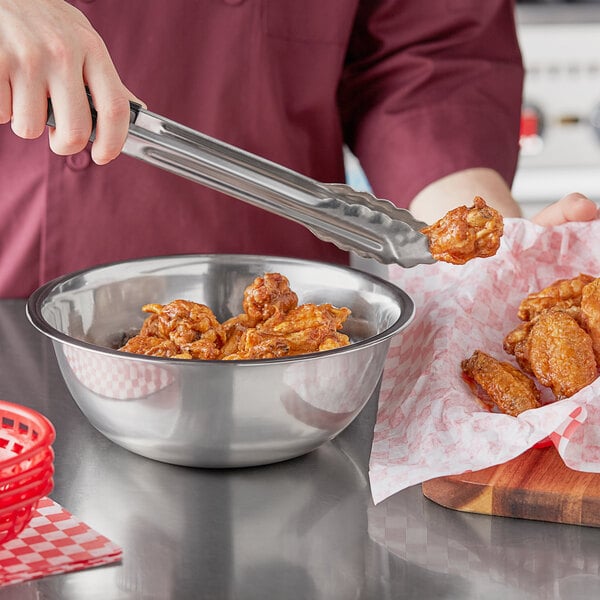 A person using tongs to cut chicken wings in a Choice stainless steel mixing bowl.