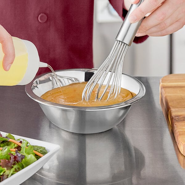 A hand using a whisk to pour salad dressing into a stainless steel mixing bowl.