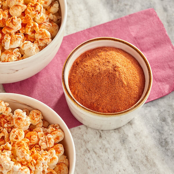 A bowl of popcorn with red and yellow powder next to a bowl of Carnival King Smokehouse Popcorn Seasoning powder.