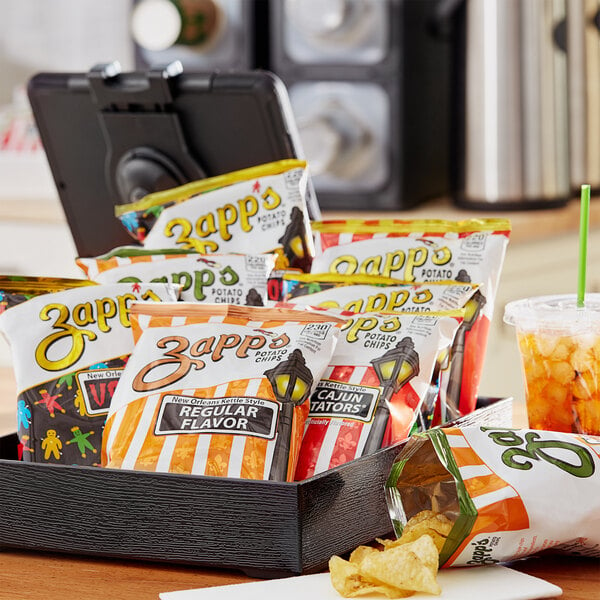 A close-up of a Zapp's Potato Chips Variety Box on a tray with a plastic cup of orange liquid.