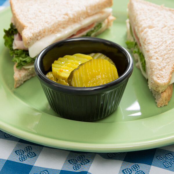 A plate with a sandwich, a ramekin of pickles, and a cup of pickles on a table in a deli.